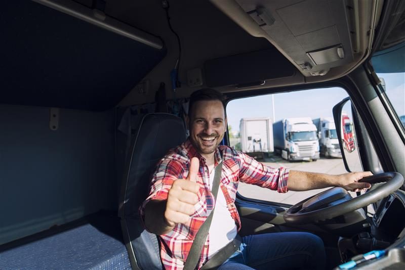 Caminhoneiro com uma mão no volante, fazendo um gesto de 'joinha' com a outra e sorrindo.