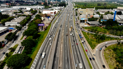 Vista aérea de rodovia com múltiplas faixas e tráfego intenso, cercada por áreas urbanas e vegetação.