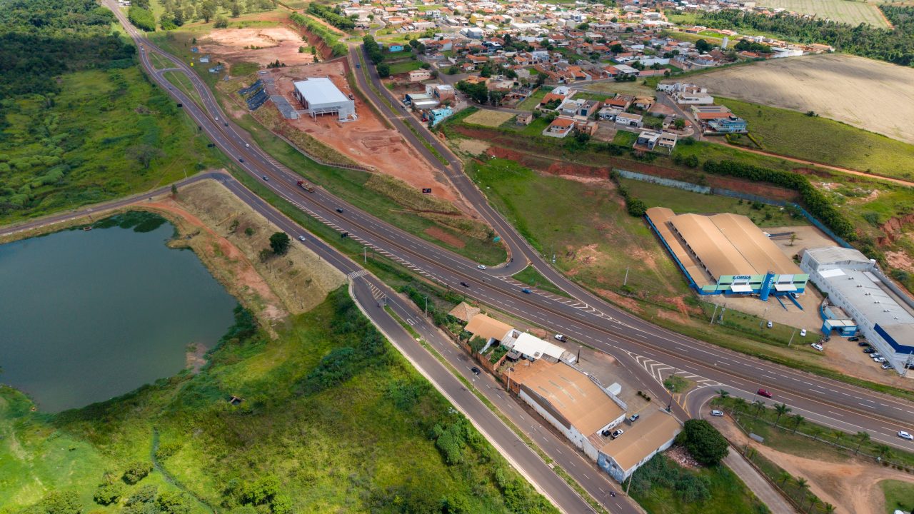Vista aérea de rodovia com entroncamento e retornos, próxima a área urbana com casas, galpões industriais e lago ao lado da via.