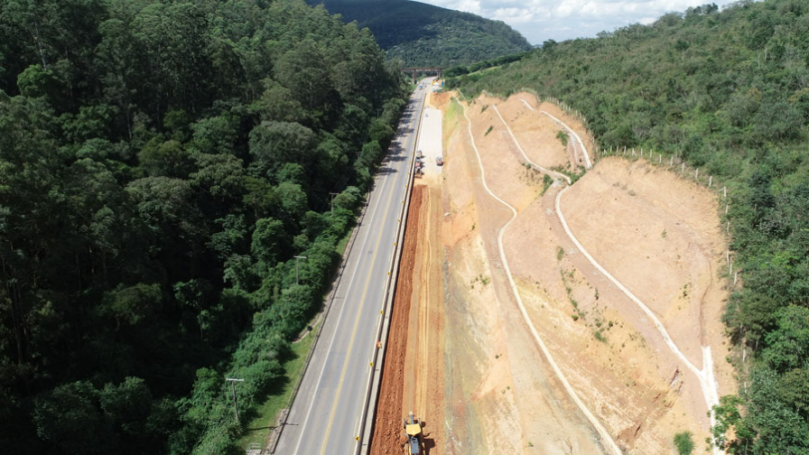 Perspectiva aérea de uma rodoviavia em meio à floresta, com obras e a infraestrutura em desenvolvimento as margens da rodovia e residências ao fundo.