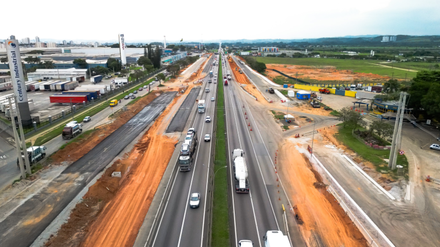 Rodovia com tráfego de veículos ao lado de construções de novas pistas marginais.