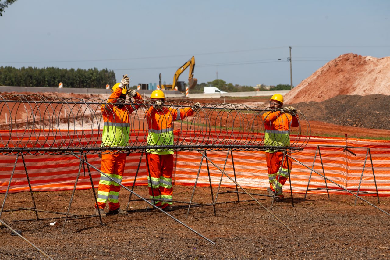 Foto de três trabalhadores da construção civil (vestindo uniformes laranja de alta visibilidade e capacetes amarelos).