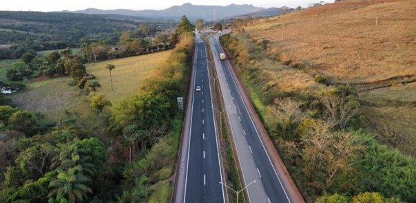 Vista aérea de uma rodovia com múltiplas faixas de tráfego próximas a edifícios industriais e armazéns, com uma paisagem urbana e colinas verdes ao fundo sob um céu parcialmente nublado.