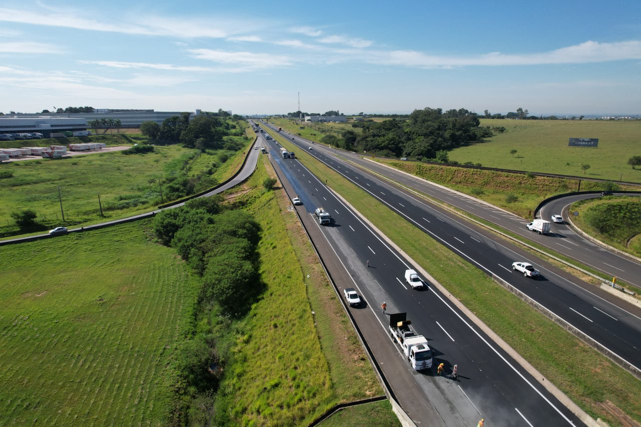 A imagem mostra a rodovia do sistema Anhanguera-Bandeirantes cercada por áreas verdes e algumas edificações industriais