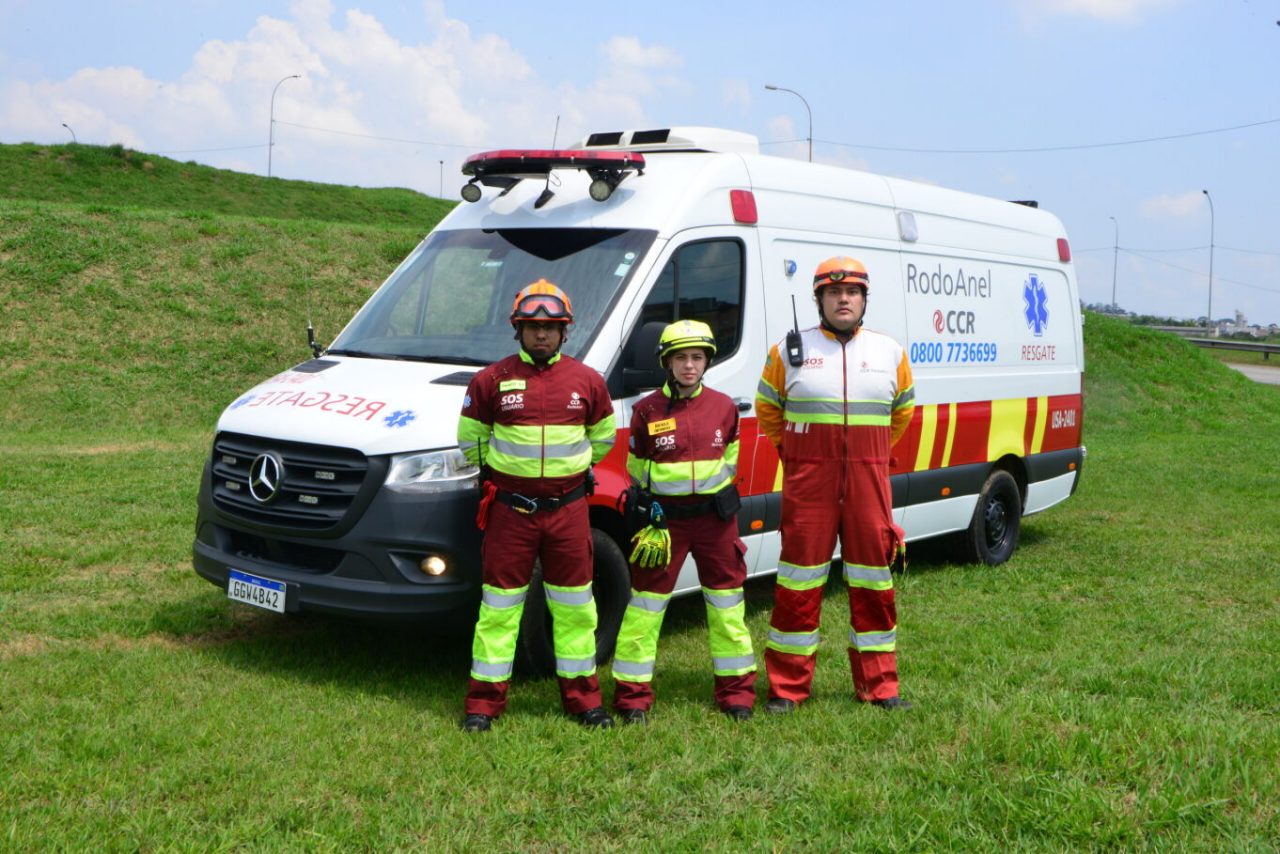 Equipe de paramédicos da CCR RodoAnel à frente da ambulância, a céu aberto e cercada por vegetação.