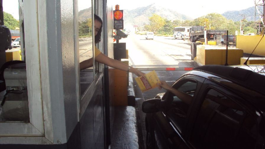 Operadora de pedágio entrega panfleto a motoristas nas rodovias, promovendo anúncios e serviços de empresas parceiras.