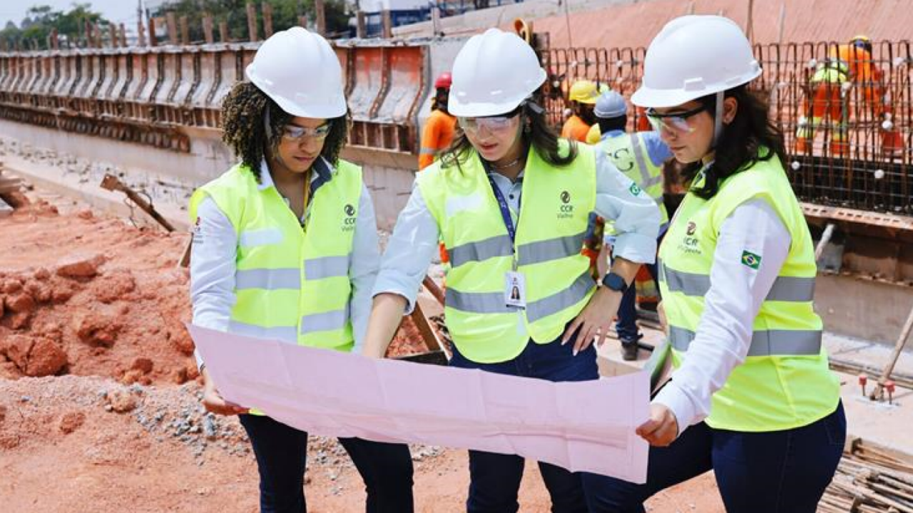 Três mulheres em vestuário de segurança analisando um plano de obra.
